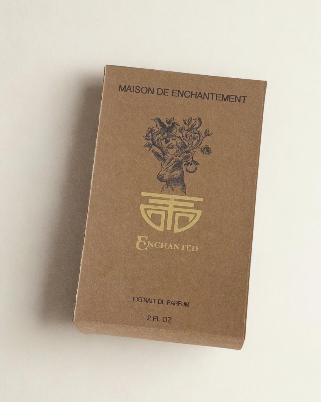 A 2 fl oz box of Maison de Enchantement Extrait de Parfum, made from brown kraft paper with gold foil lettering and a deer illustration, set against a plain off-white background.
