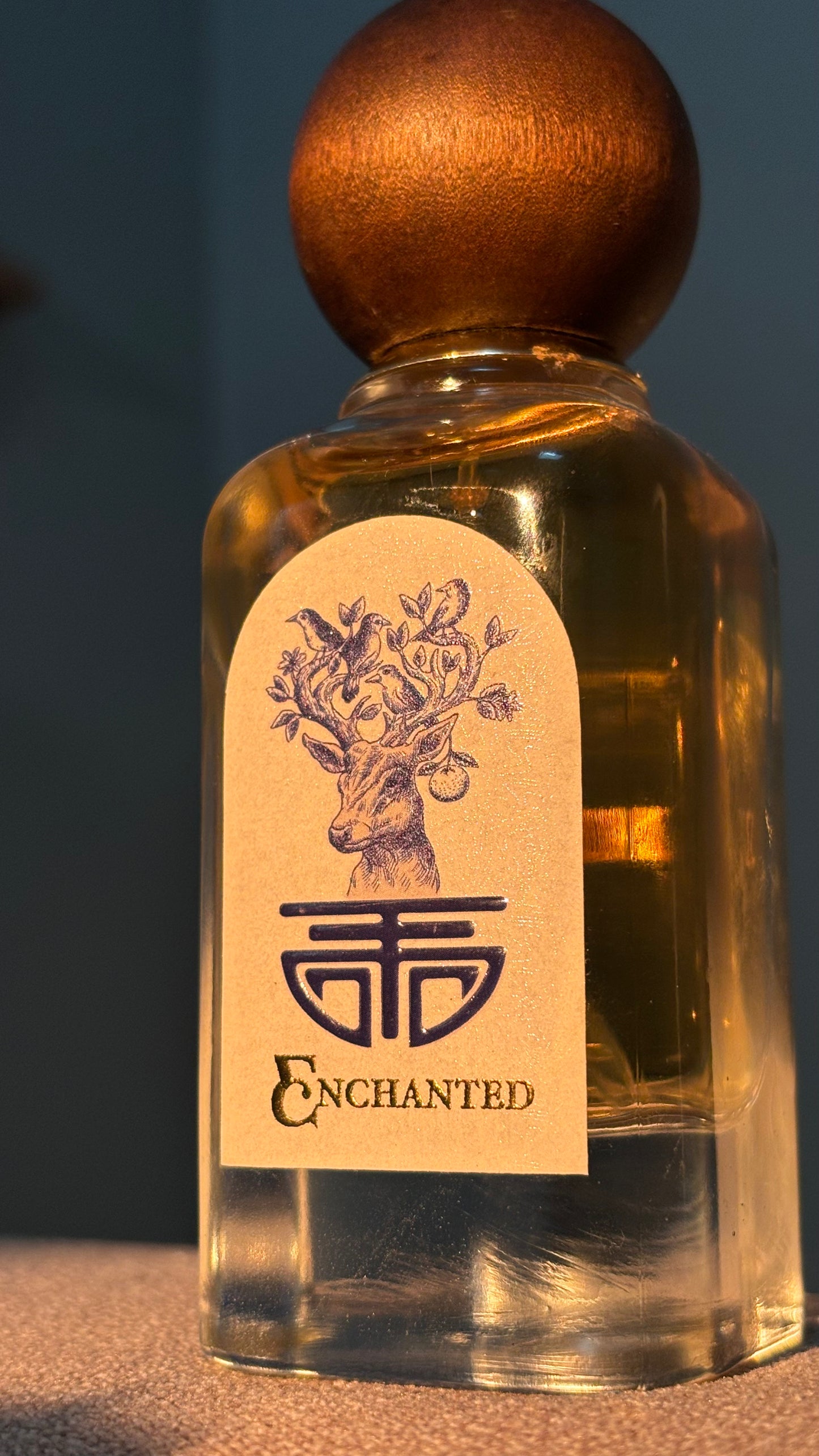 A clear glass perfume bottle filled with amber liquid, topped with a round metallic gold cap, and featuring a cream label with a deer illustration and the word 'ENCHANTED'.